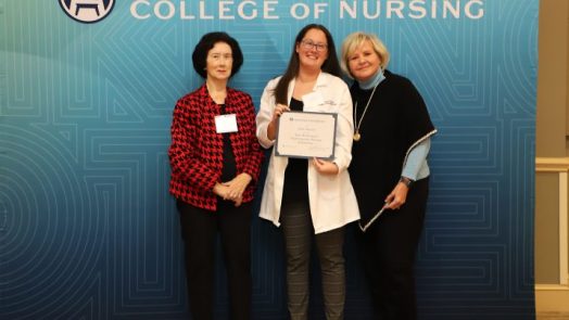 Three women stand together, the middle one holding up a certificate.