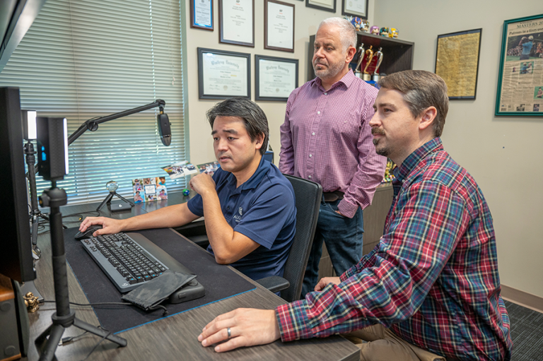 Two men sit and one man stands around a computer in an office.