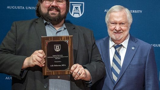 Two men stand together as one holds up an award.