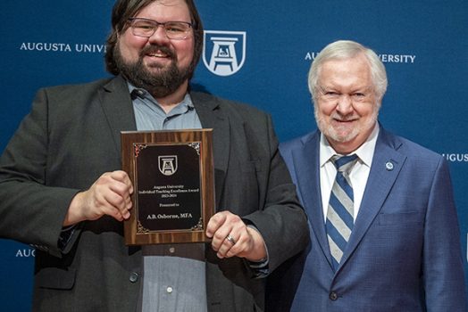 Two men stand together as one holds up an award.