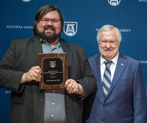 A man holding a plaque stands next to another man, both smiling for the camera in front of a navy blue Augusta University backdrop.