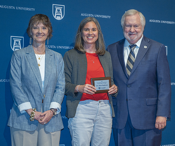 Two women and a man smile at the camera in front of a navy Augusta University backdrop. The woman in the middle holds the plaque she received.
