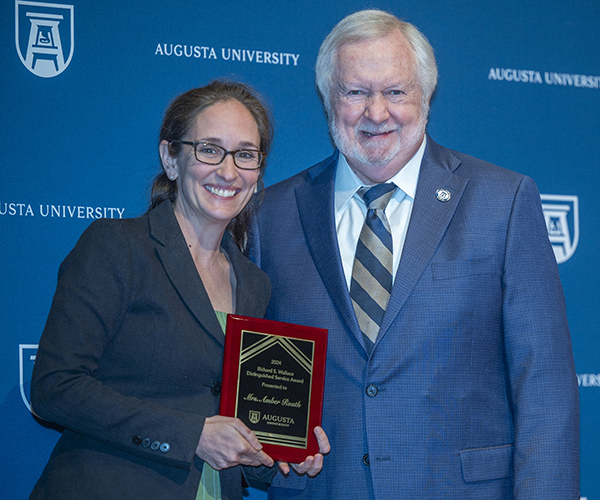 A woman holding a plaque stands next to a man, both smiling for the camera in front of a navy blue Augusta University backdrop.