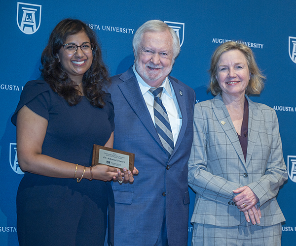 Two women stand on either side of a  man, smiling at the camera in front of a navy Augusta University backdrop. The woman on the left holds the plaque she received.
