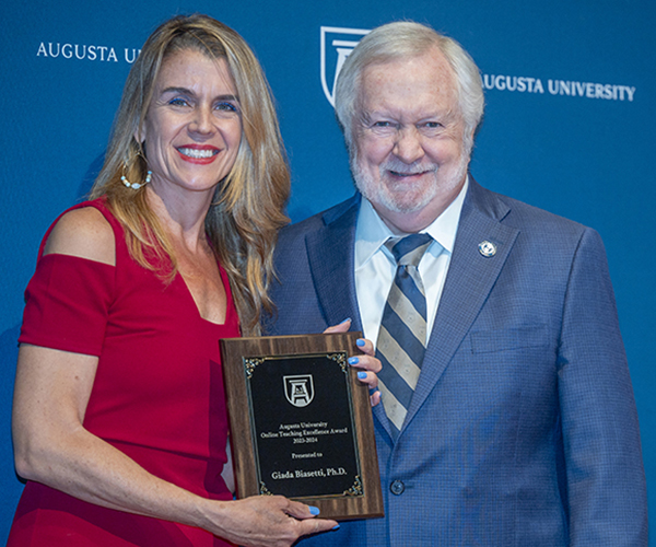 A woman holding a plaque stands next to a man, both smiling for the camera in front of a navy blue Augusta University backdrop.
