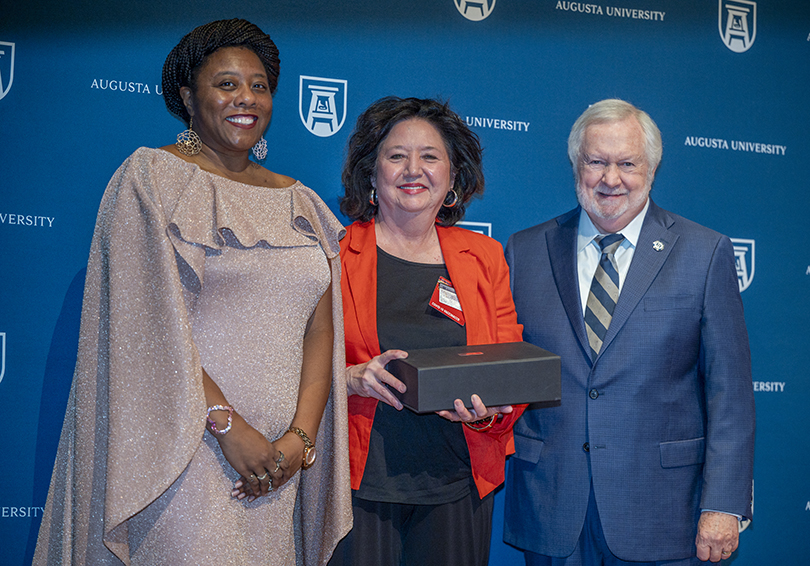 Two woman and a man smile at the camera. The woman in the middle holds a box containing her award.