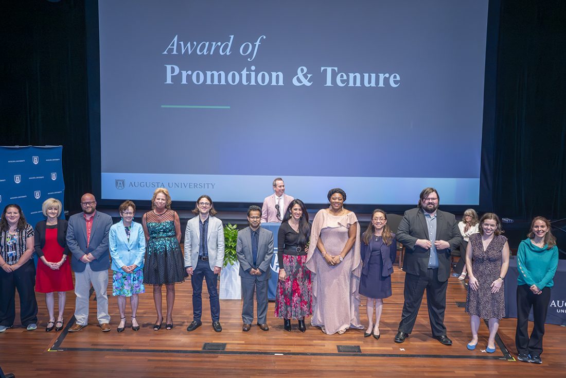 Thirteen men and women stand in a line on a stage with the words Award of Promotion and Tenure on a large screen behind them
