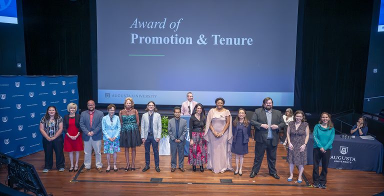 Thirteen men and women stand in a line on a stage with the words Award of Promotion and Tenure on a large screen behind them