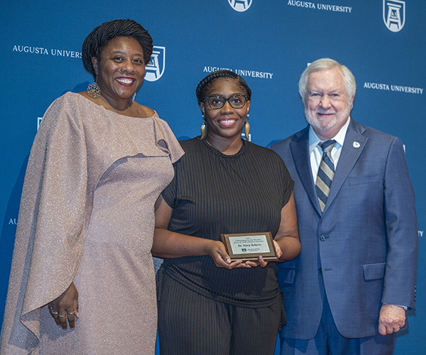 Two women and a man smile at the camera in front of a navy Augusta University backdrop. The woman in the middle holds the plaque she received.