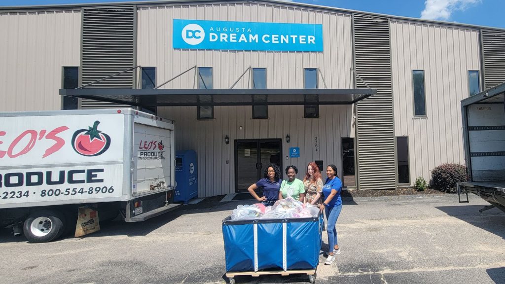 Four women stand together in front of the Augusta Dream Center with a large cart filled with bags of clothing.
