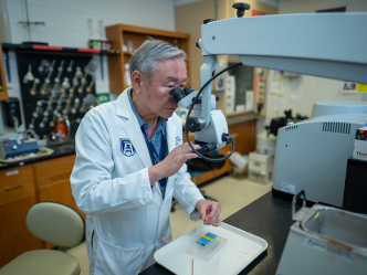 A male scientist wearing a lab coat looks into a large microscope at slides on a table.