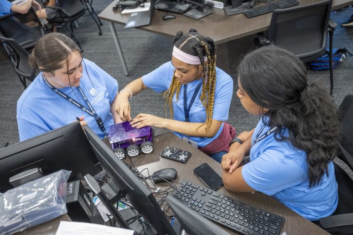 Three high school girls work on a robot with wheels during a camp at a university.
