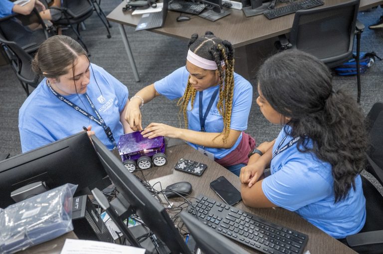 Three high school girls work on a robot with wheels during a camp at a university.