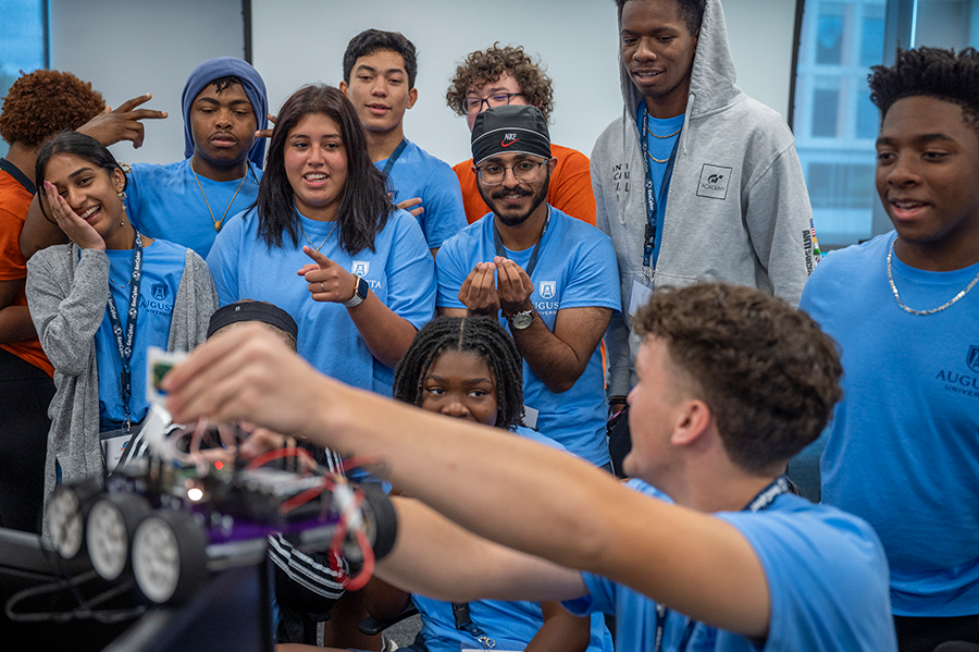 A group of male and female high school students laugh and instruct a friend on where to place a remote-control car on top of several computer screens.