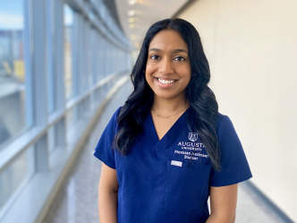 A woman stands in a long hallway next to a wall of windows. She is smiling and wearing medical scrubs that have "Augusta University Physician Assistant Student" embroidered over the front pocket.