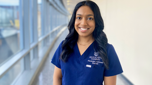 A woman stands in a long hallway next to a wall of windows. She is smiling and wearing medical scrubs that have "Augusta University Physician Assistant Student" embroidered over the front pocket.