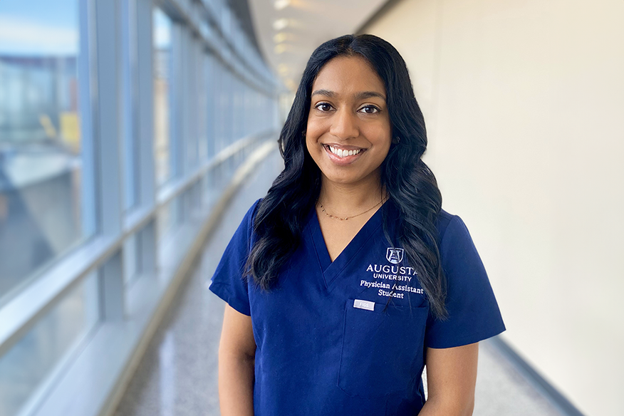 A woman stands in a long hallway next to a wall of windows. She is smiling and wearing medical scrubs that have "Augusta University Physician Assistant Student" embroidered over the front pocket.