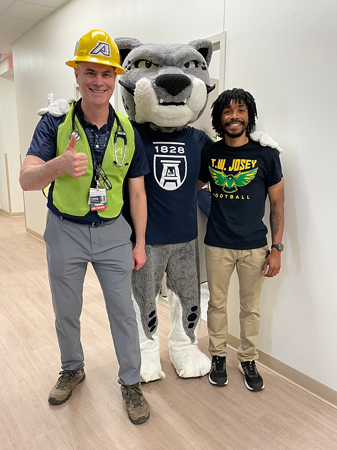 Two men stand with a collegiate sports mascot. The mascot is a jaguar or large cat wearing a t-shirt. One of the men is wearing a construction hardhat and safety vest.