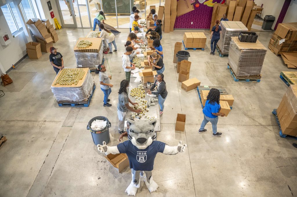 Aerial shot of college students volunteering at a food bank while Augustus the mascot stands in front with his arms wide open.