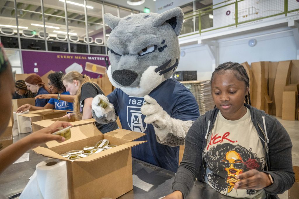 A row of students package food containers at a food bank with help from Augustus the mascot.