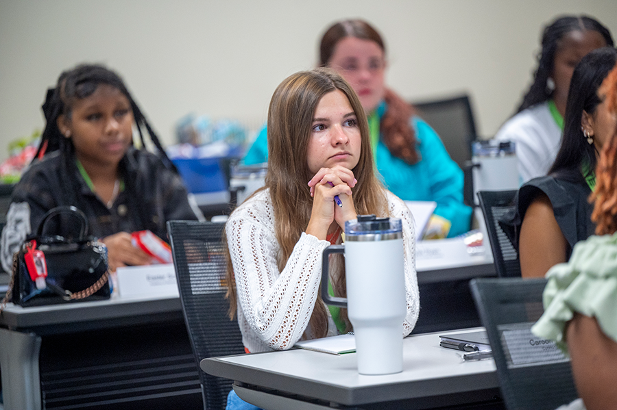 A high school girl sits at a long table with other girls in a college classroom.