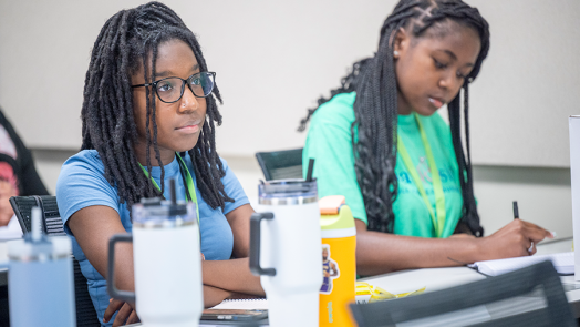 Two female high school students sit a a long desk and take notes during a lecture.