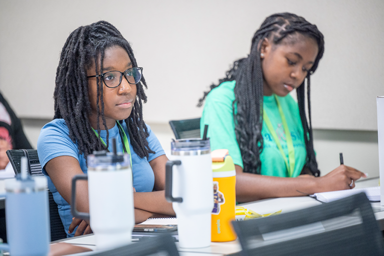 Two female high school students sit a a long desk and take notes during a lecture.