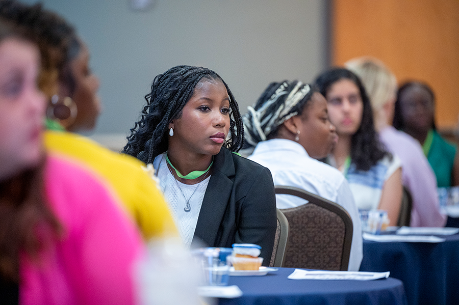 A high school girl sits at a table surrounded by other girls while listening to a lecture given by a college professor.