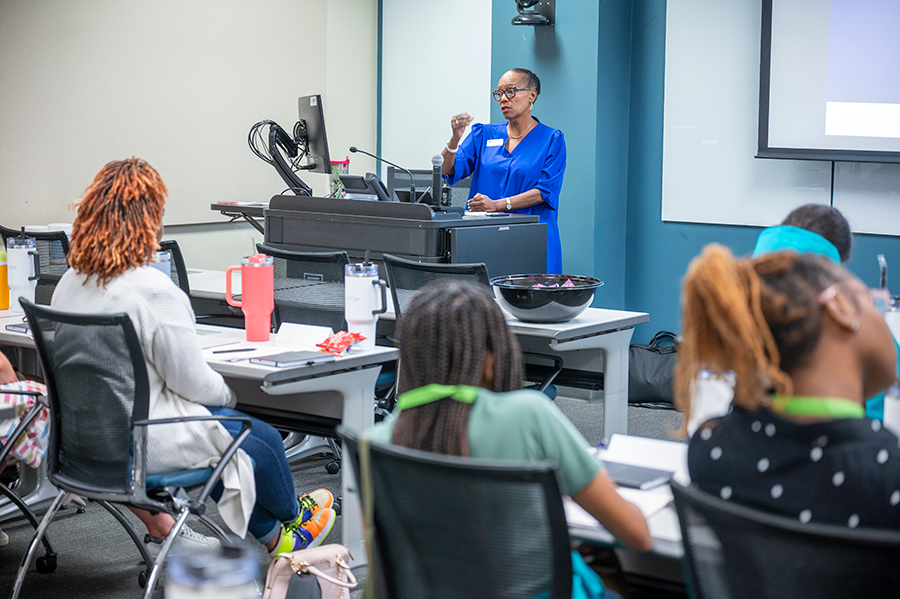 A woman stands at a computer at the front of the room, talking to a classroom full of high school girls