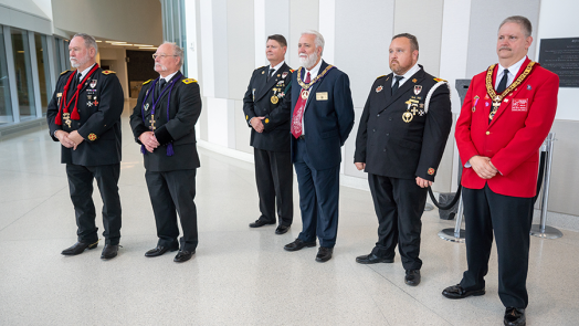 Six men in suits stand in a large atrium of a building. Each has numerous medals pinned to his chest.