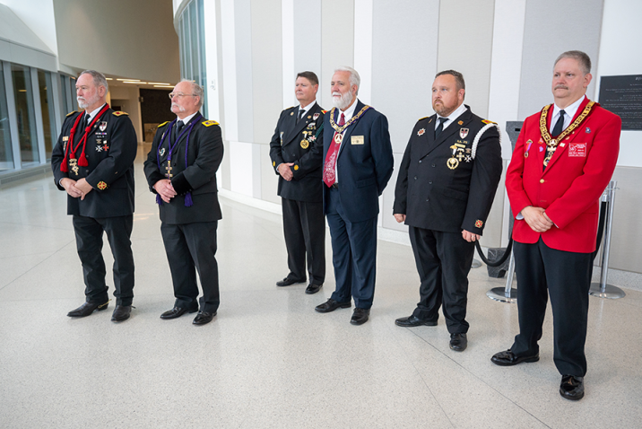 Six men in suits stand in a large atrium of a building. Each has numerous medals pinned to his chest.