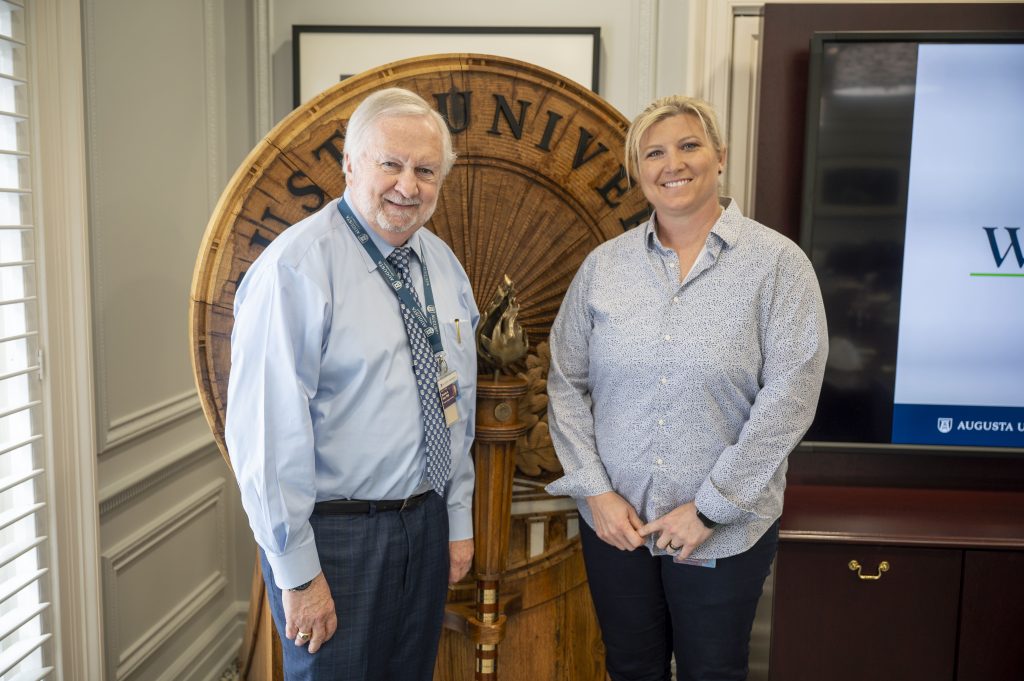 A man and woman pose for a photo in front of the Augusta University shield.