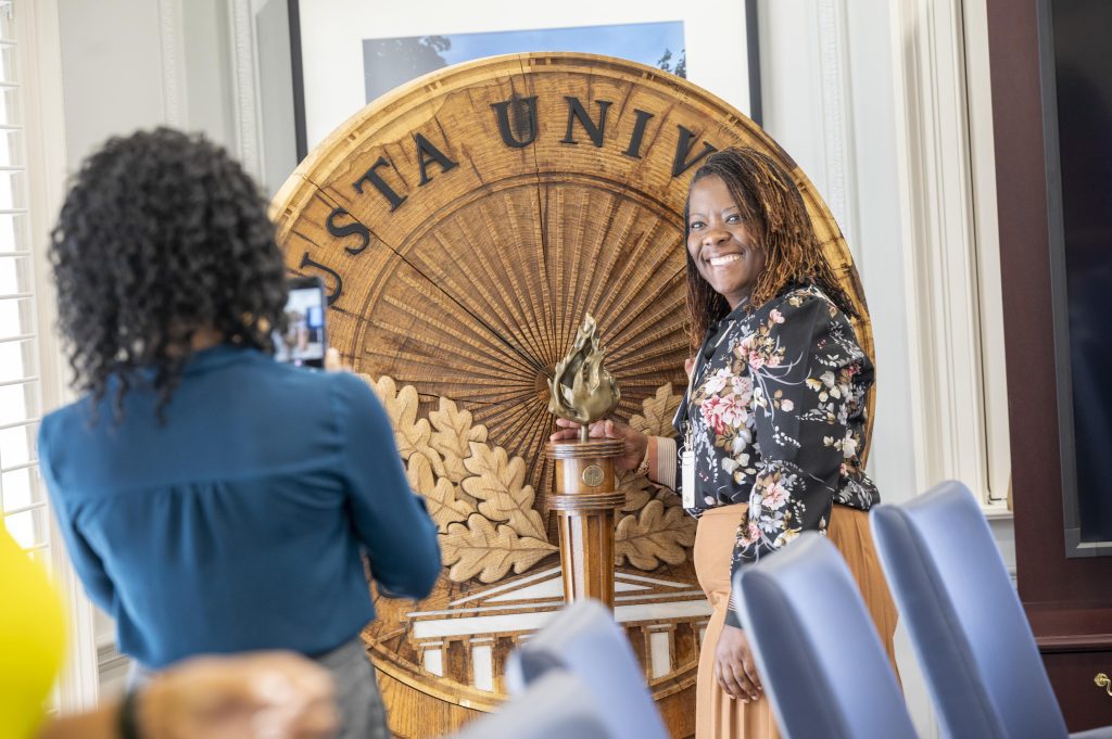 A woman poses in front of the Augusta University shield while another woman takes a photo of her.