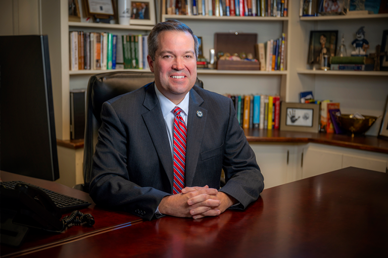 A man sits at a desk inside an office wearing a suit and tie. There are books on the shelves behind him and a computer sitting on the desk off to one side.