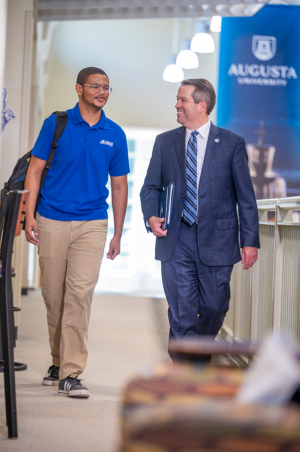 A man in a suit walks next to and talks to a college student in a hallway.