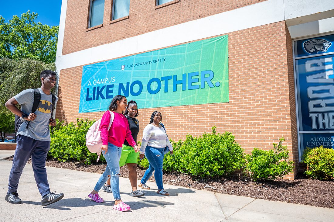 four students walk on the sidewalk in front of a building with a sign that reads "Like No Other"