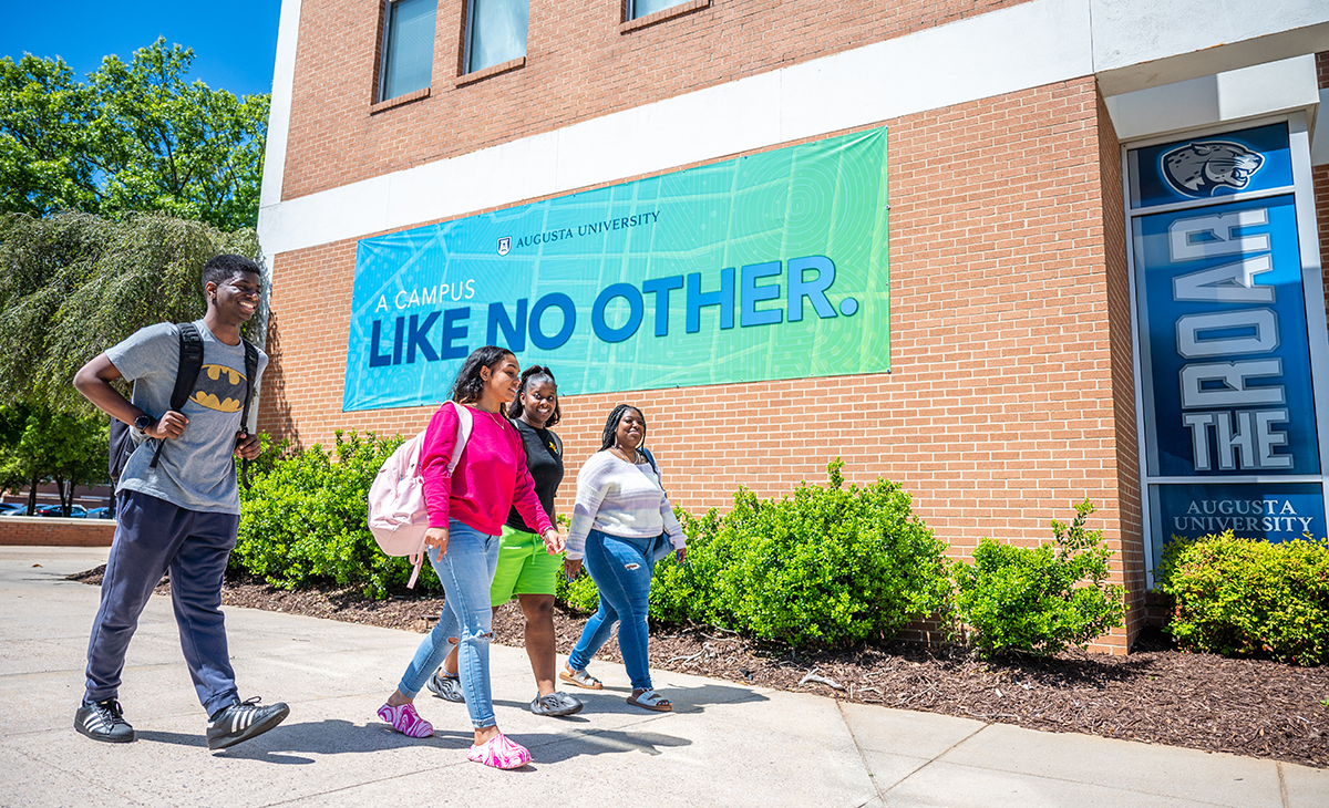 four students walk on the sidewalk in front of a building with a sign that reads "Like No Other"