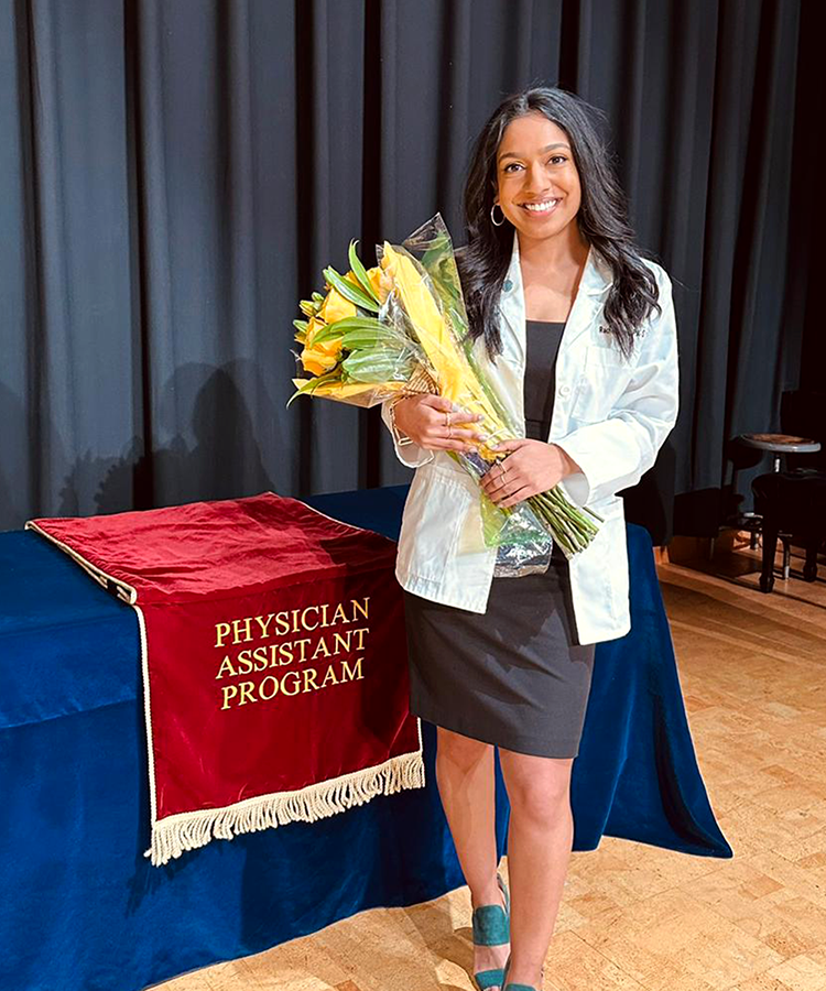 A young woman wears a medical professional's coat while holding a bouquet of flowers on a stage. There is a table behind her with a dark red flag draped over it that has "Physician Assistant Program" embroidered on it.