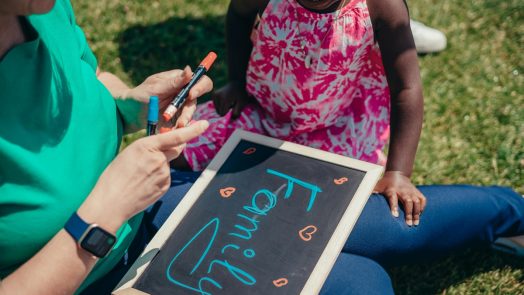 A young child sits outside in the grass with a woman holding a chalkboard that says family