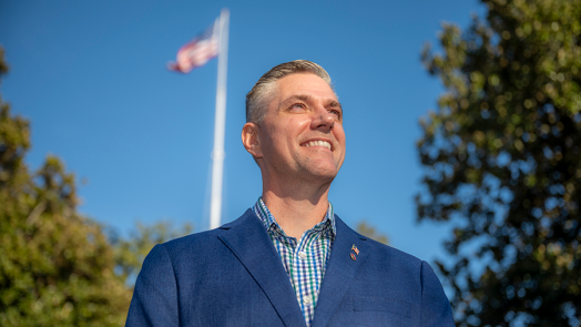 A man in a suit stands outside with the American Flag behind him.