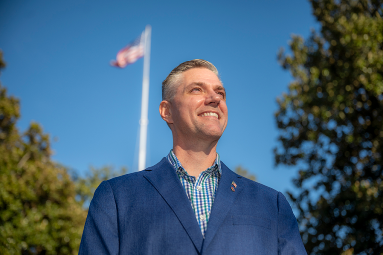 A man in a suit stands outside with the American Flag behind him.