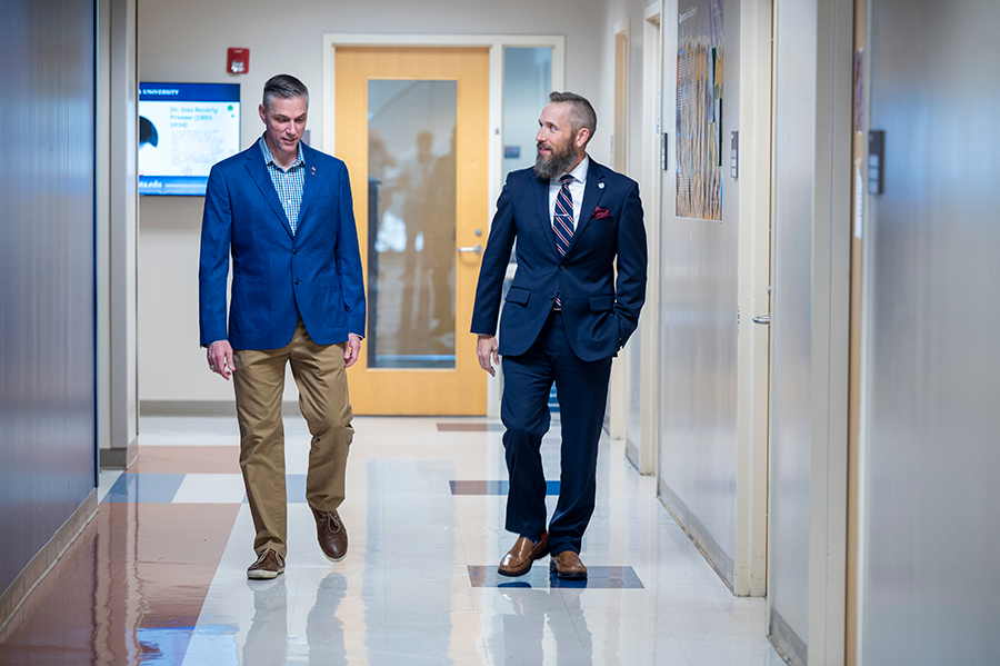 Two men in suits walk down a hallway while talking to each other.