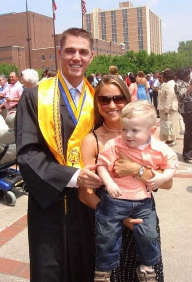 A man wearing college graduation gown and stole stands next to a woman holding a toddler.