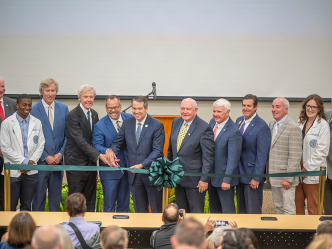 A large group of people stand at the front of a big auditorium. Three men in the middle of the group are holding a ceremonial pair of over-sized scissors to cut a ribbon signifying the opening of a new college campus.