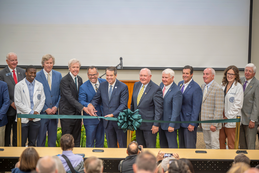 A large group of people stand at the front of a big auditorium. Three men in the middle of the group are holding a ceremonial pair of over-sized scissors to cut a ribbon signifying the opening of a new college campus.