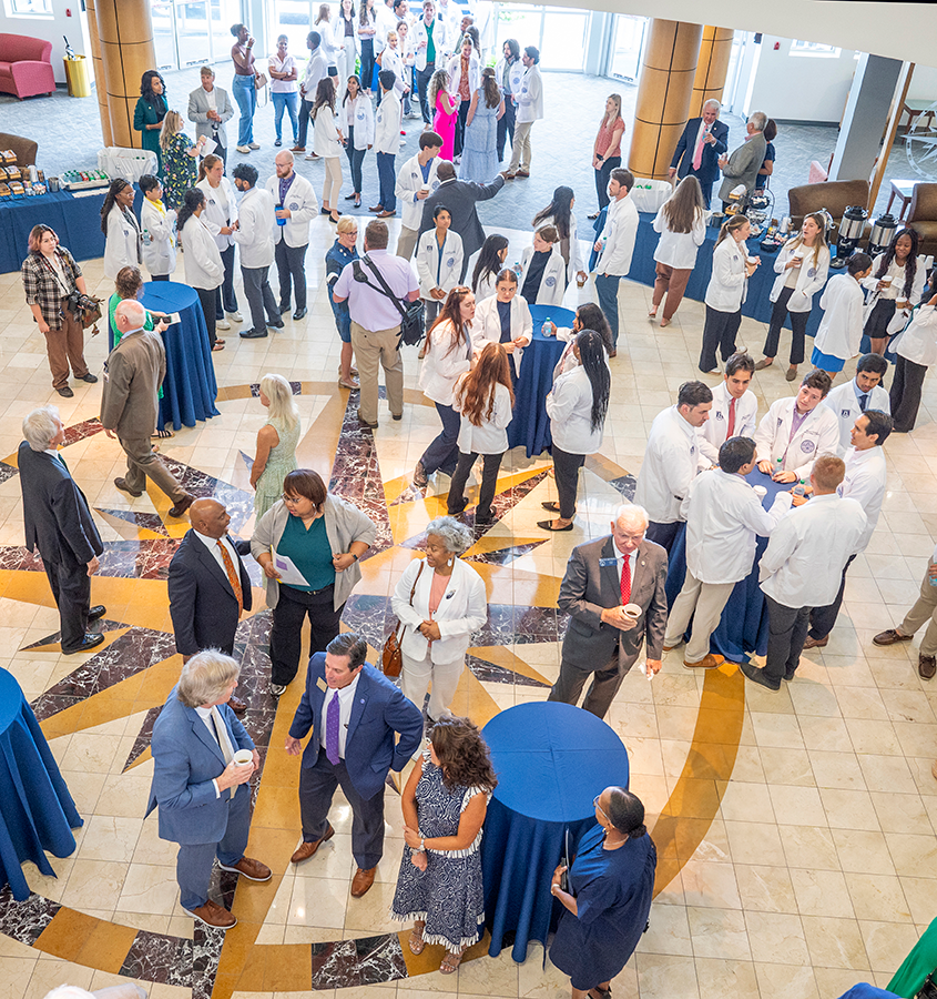 People stand around a large atrium during a special event. Some are holding cups of coffee or juice. Many of those gathered are wearing lab coats for medical professionals.