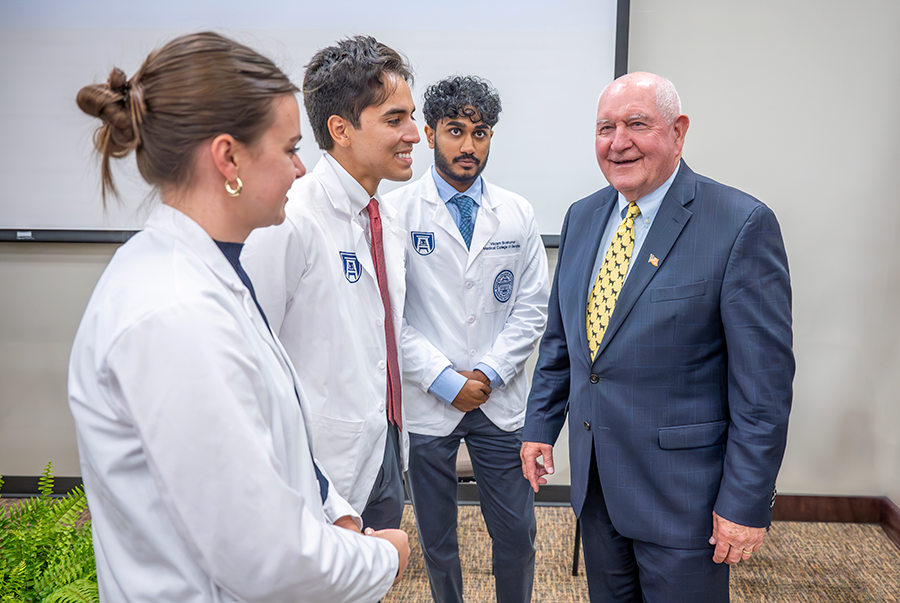 An older man in a suit talks with three medical students, all wearing lab coats, during a special ceremony.