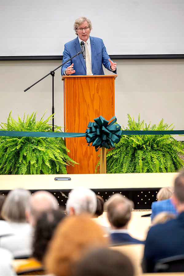 A man in a suit and tie stands at a podium with a microphone in front of a large auditorium that is filled with people.