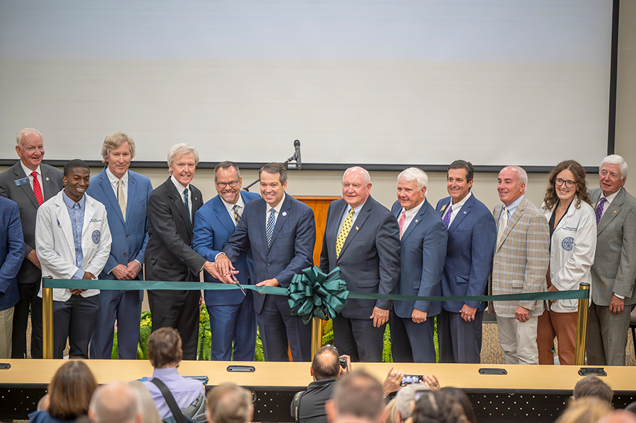 A large group of people stand at the front of a big auditorium. Three men in the middle of the group are holding a ceremonial pair of over-sized scissors to cut a ribbon signifying the opening of a new college campus.