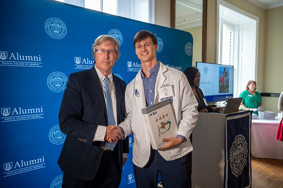 A man and a college-aged male student wearing a medical professional's lab coat stand in front of a large room. The young man holds a box that holds a stethoscope inside.